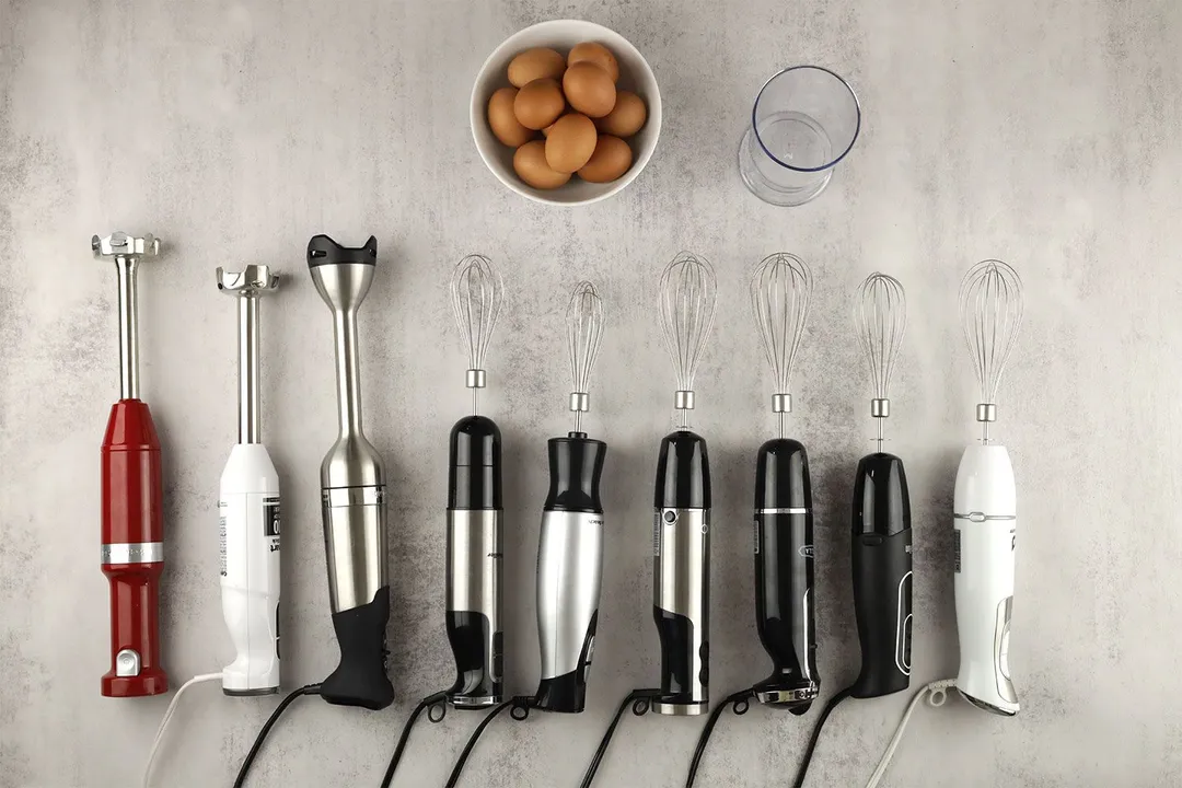 Nine immersion blenders lying on a table with ingredients and necessary tools for the whipped egg-white test, including eggs contained in a white bowl and a plastic beaker, next to them.