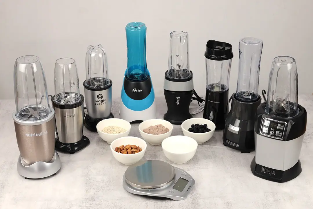 8 personal blenders on a table ready for their protein shake test, with five bowls containing five different ingredients and a scale next to them. 