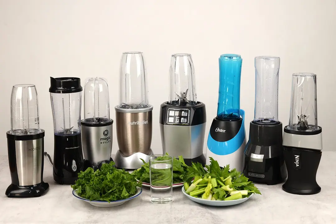8 personal blenders on a table ready for their test, with a glass of water and three plates of fibrous greens next to them.
