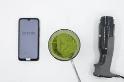 A plastic beaker containing a batch of green smoothie whose parts are scooped with a stainless steel spoon is between the Breville’s motor body and a smartphone displaying the total blending time (2 minutes and 21 seconds). 