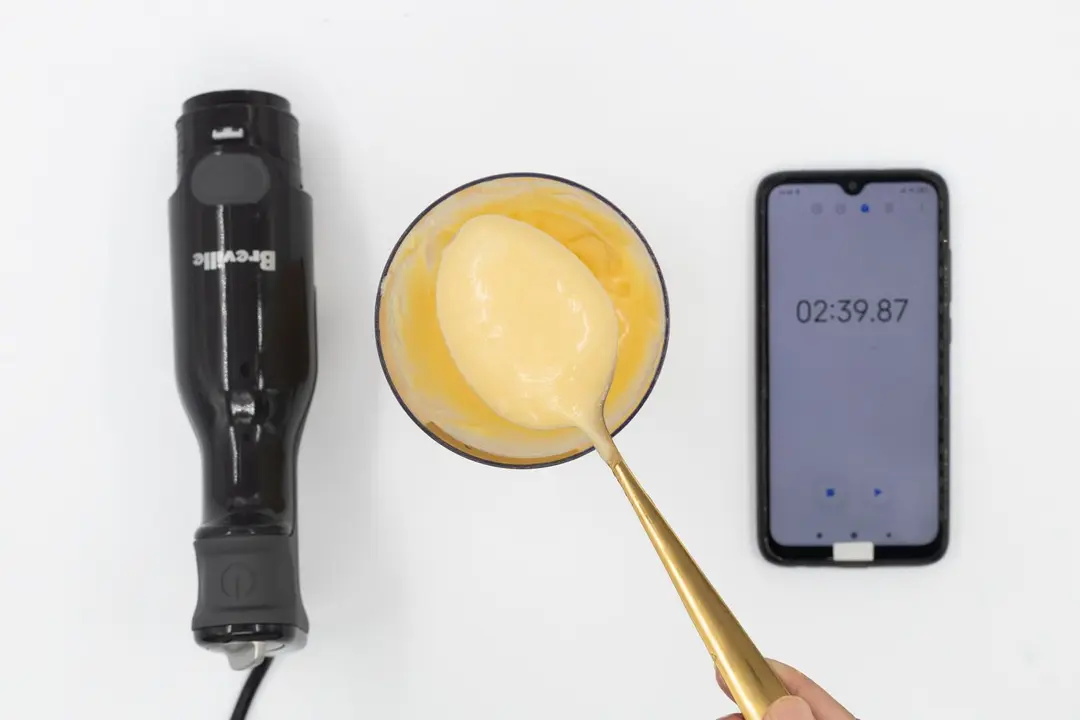 Scooping a spoon of mayonnaise from its full batch made by the Breville blender and contained in a plastic beaker that stands between the Breville’s motor and a smartphone displaying the total mixing time ( 2 minutes and 39 seconds).