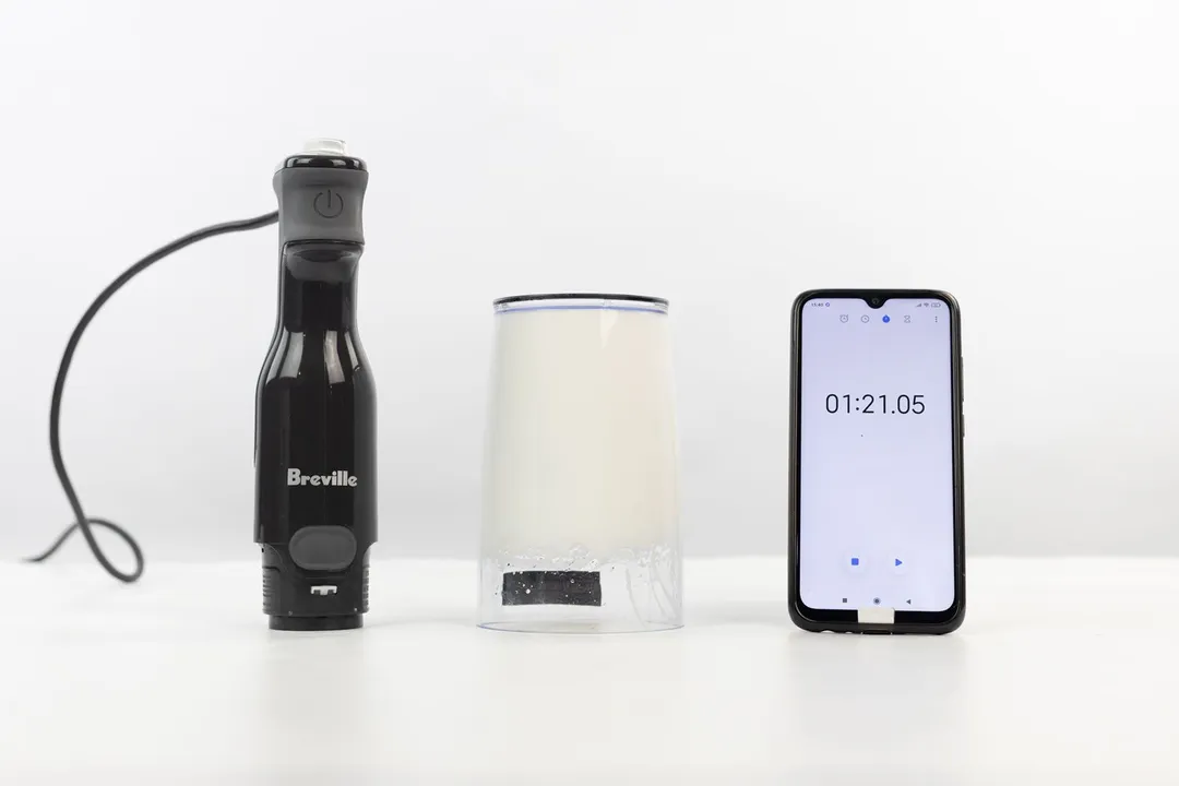 The plastic beaker containing testing beaten egg-white of the Breville immersion blender is put upside down on the white table with its motor body and a smartphone displaying the total whipping time (1 minute and 21 seconds) by its sides.