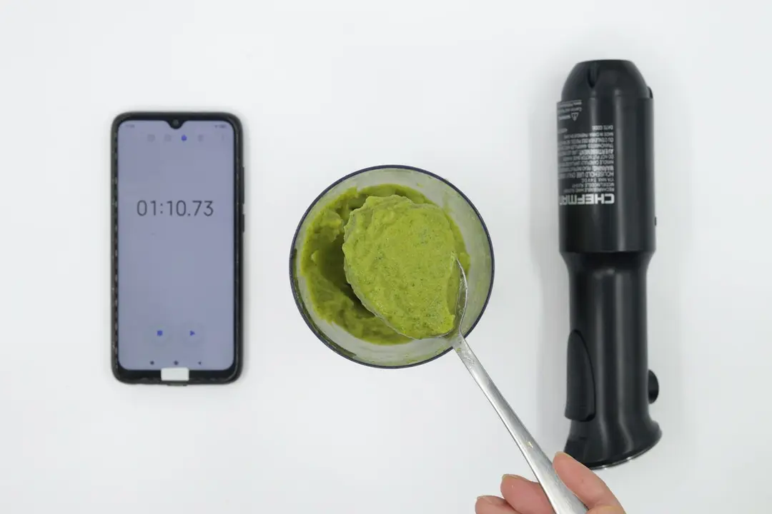 A plastic beaker containing a batch of green smoothie whose parts are scooped with a stainless steel spoon is between the Chefman’s motor body and a smartphone displaying the total blending time (1 minute and 10 seconds).