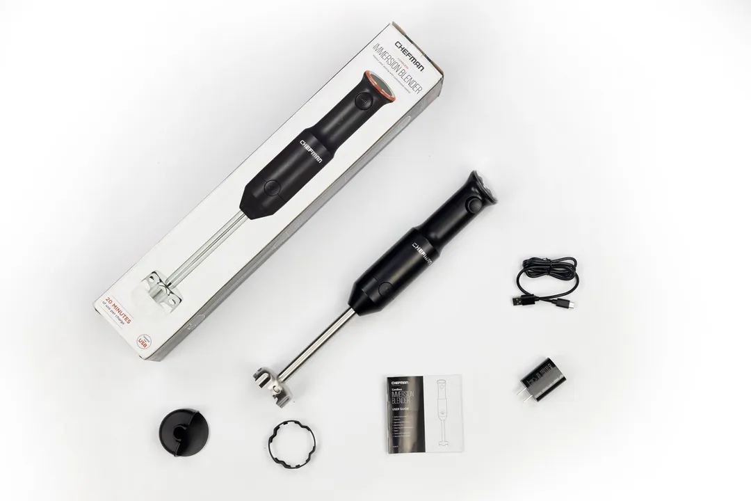 An owner manual, USB cord, charger adapter, immersion blending wand with its motor body attached, blade edge guard, blade guard, and paper cartoon box of The Chefman cordless hand blender being side by side on a white table.