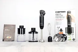 Unboxing the Chefman Immersion Blender, from left to right: a user manual, a spiralizer, a food processor, a blending wand with its motor body attached, a whisk attachment, three spiralizing blades, and a paper carton box.