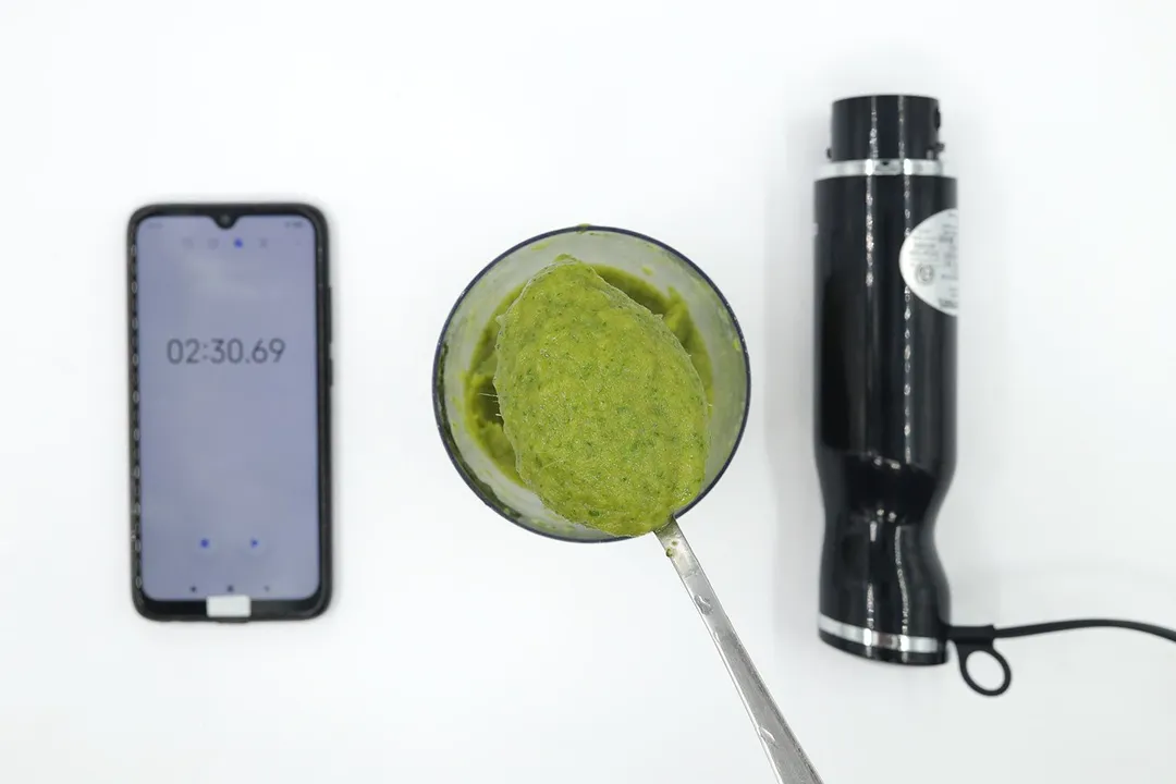 A plastic beaker containing a batch of green smoothie whose parts are scooped with a stainless steel spoon is between the Chefman Vegetable Slicer 6-in-1 Immersion Blender motor body and a smartphone displaying the total blending time (2 minutes and 30 seconds).