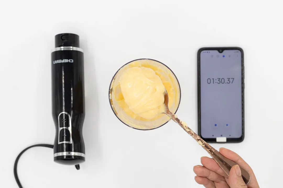 Scooping a spoon of mayonnaise from its full batch made by the Chefman immersion blender and contained in a plastic beaker that stands between the Chefman’s motor and a smartphone displaying the total mixing time ( 1 minute and 30 seconds).
