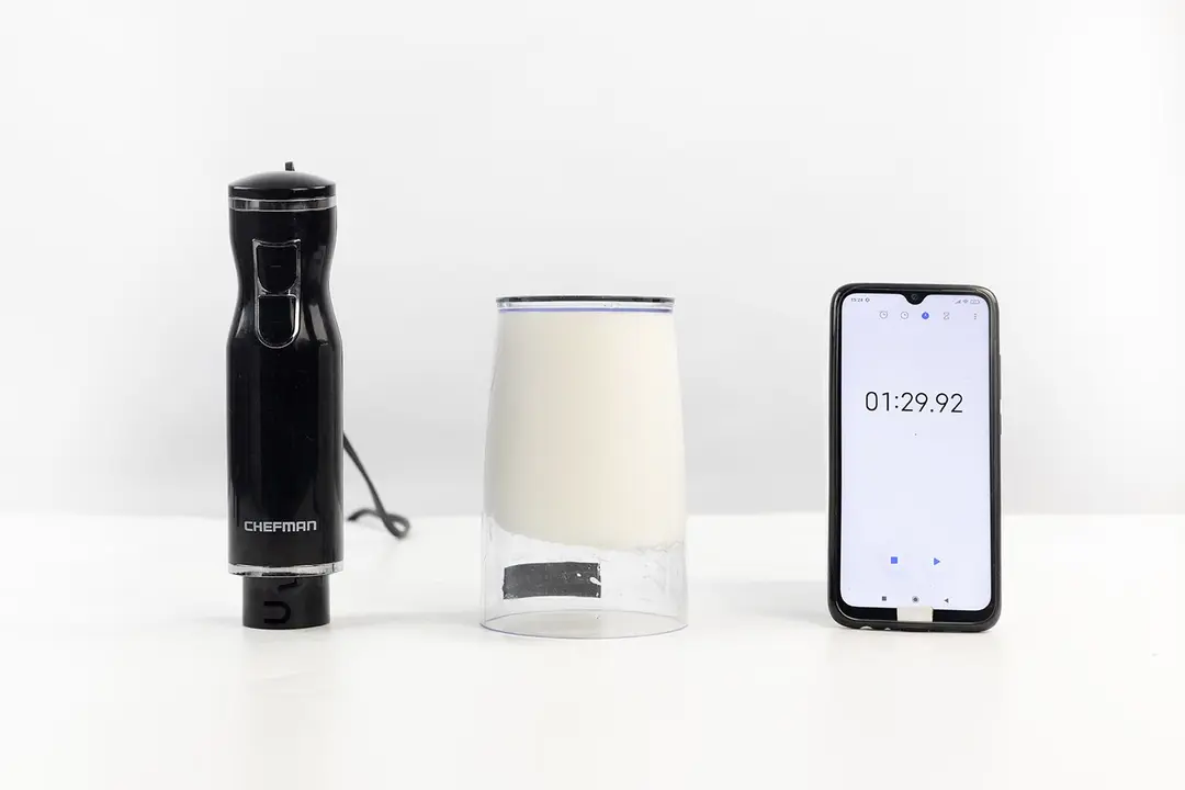The plastic beaker containing testing beaten egg-white of the Chefman immersion blender is put upside down on the white table with its motor body and a smartphone displaying the total whipping time (1 minute and 29seconds) by its sides.