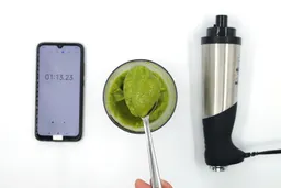 A plastic beaker containing a batch of green smoothie whose parts are scooped with a stainless steel spoon is between the All-Clad’s motor body and a smartphone displaying the total blending time (1 minute and 13 seconds).