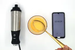 Scooping a spoon of failed mayonnaise made by the All-Clad hand blender from its full batch in a plastic beaker which is standing between the All-Clad’s motor body and a smartphone displaying the total mixing time (2 minutes and 5 seconds). 