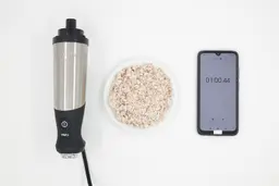A white plate of almond pulp being between the All-Clad’s motor body and a smartphone displaying the total grinding time (1 minute).