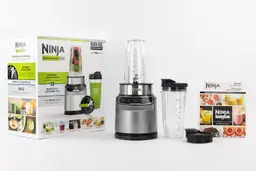The Ninja BN401 Nutri Pro Personal Blender and its additional accessories by its side, including an extra blending cup with lid, a spout lid, a paper carton box, and a user guide.