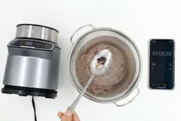 A batch of protein shake prepared by the Ninja BN401 Nutri Pro Single-Serve Blender is checked for smoothness by being drained through a stainless steel mesh strainer, with a smartphone displaying the total blending time (1 minute) next to it.