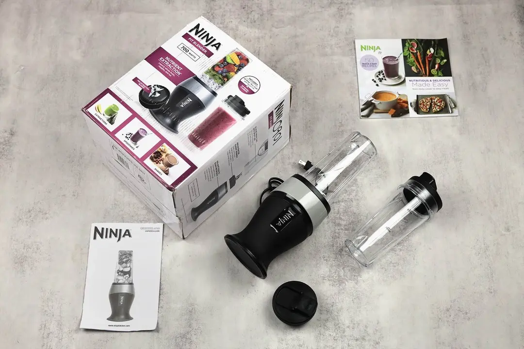 Unboxing the Ninja QB3001SS Fit personal blender, from left to right: a paper carton box, a to-go lid, the unit’s motor base with its blending cup and blade assembly attached, an additional blending cup with to-go lid, and a user’s guide.