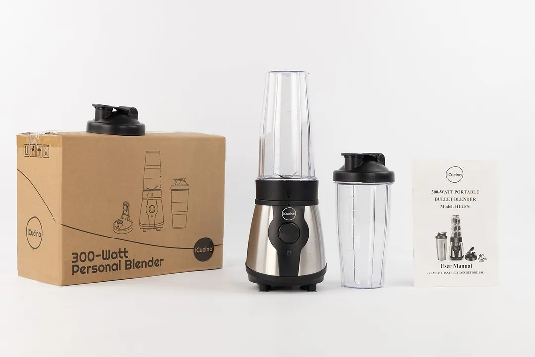 The iCucina Personal Blender standing on a white table with its accessories, including a to-go lid, an extra blending cup with lid, a user’s manual, and a paper carton box, by its sides.