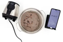 A batch of protein shake packed with dried blueberries, oatmeal, and almonds prepared by the iCucina Personal Blender is checked for smoothness by being drained through a stainless steel mesh strainer, with a smartphone displaying the total blending time (1 minute and 49 seconds) next to it.