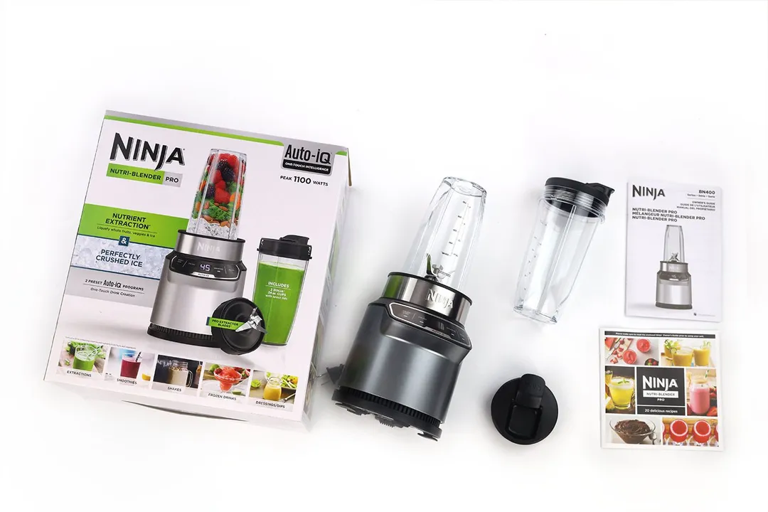 The Ninja BN401 Nutri Pro personal blender lying on a white table with the accessories by its side, including a blending cup with lid, a spout lid, a paper carton box, a recipe book, and owner’s manuals.