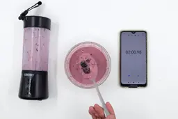 A spoon of fruity smoothie packed with blueberries, blackberries, strawberries, and mango made by the OBERLY portable blender with a smartphone displaying the total blending time ( 1 minute and 40 seconds) next to it.