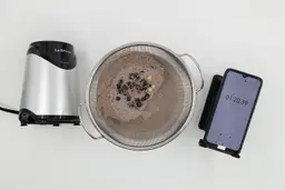 A batch of protein shake prepared by the La Reveuse is checked for smoothness by being drained through a stainless steel mesh strainer, with a smartphone displaying the total blending time (1 minute and 20 seconds) next to it.