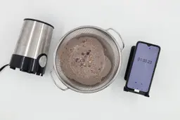 A batch of protein shake prepared by the BELLA Single-Serve Blender is checked for smoothness by being drained through a stainless steel mesh strainer, with a smartphone displaying the total blending time (1 minutes and 30 seconds) next toit.