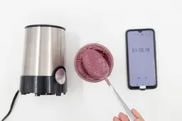 A spoon of fruity smoothie packed with blueberries, blackberries, strawberries, and mango made by the BELLA portable blender with a smartphone displaying the total blending time ( 1 minute and 10 seconds) next to it.