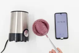 A spoon of fruity smoothie packed with blueberries, blackberries, strawberries, and mango made by the BELLA portable blender with a smartphone displaying the total blending time ( 1 minute and 10 seconds) next to it.