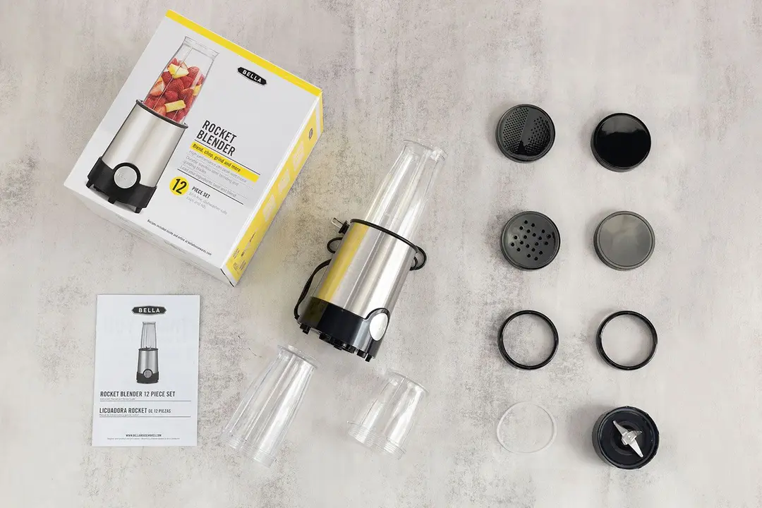 The BELLA personal blender lying on a table with its additional accessories, including two blending cups, two storage lids, two shaker lids, a blade assembly, two lip rings, a user guide, and a paper carton box.