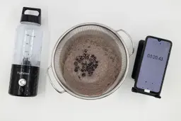 A batch of protein shake prepared by the PopBabies Single-Serve Blender is checked for smoothness by being drained through a stainless steel mesh strainer, with a smartphone displaying the total blending time (3 minutes and 20 seconds) next to it.