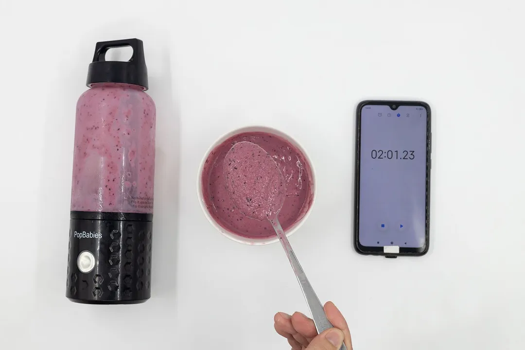 A spoon of fruity smoothie packed with blueberries, blackberries, strawberries, and mango made by the PopBabies  portable blender with a smartphone displaying the total blending time ( 2 minutes and 01 seconds) next to it.