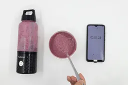 A spoon of fruity smoothie packed with blueberries, blackberries, strawberries, and mango made by the PopBabies  portable blender with a smartphone displaying the total blending time ( 2 minutes and 01 seconds) next to it.