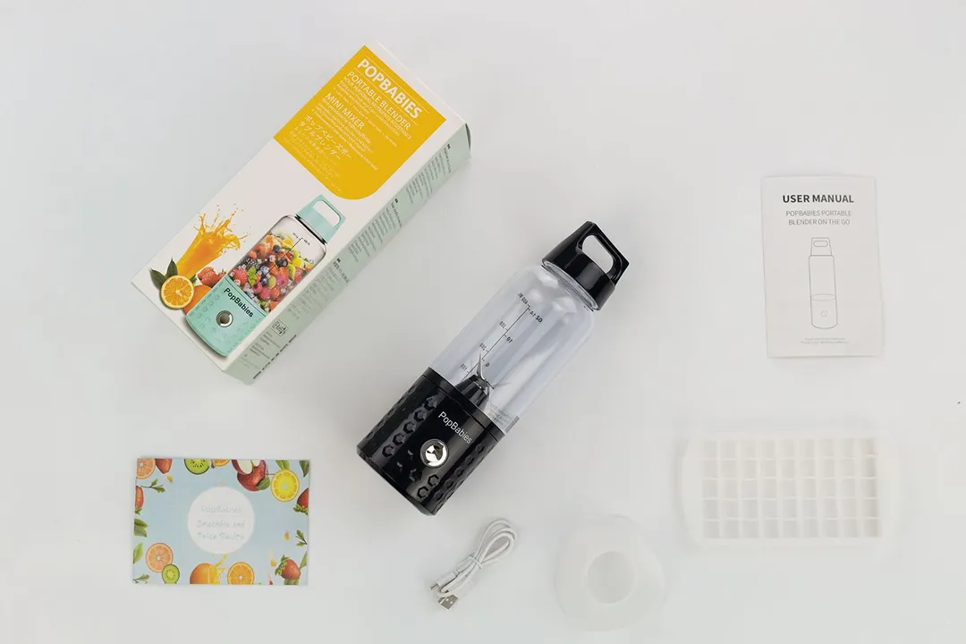 The PopBabies  personal blender lying on a table with a paper carton box, user guide, charging cable, funnel, and ice cube tray by its sides. 
