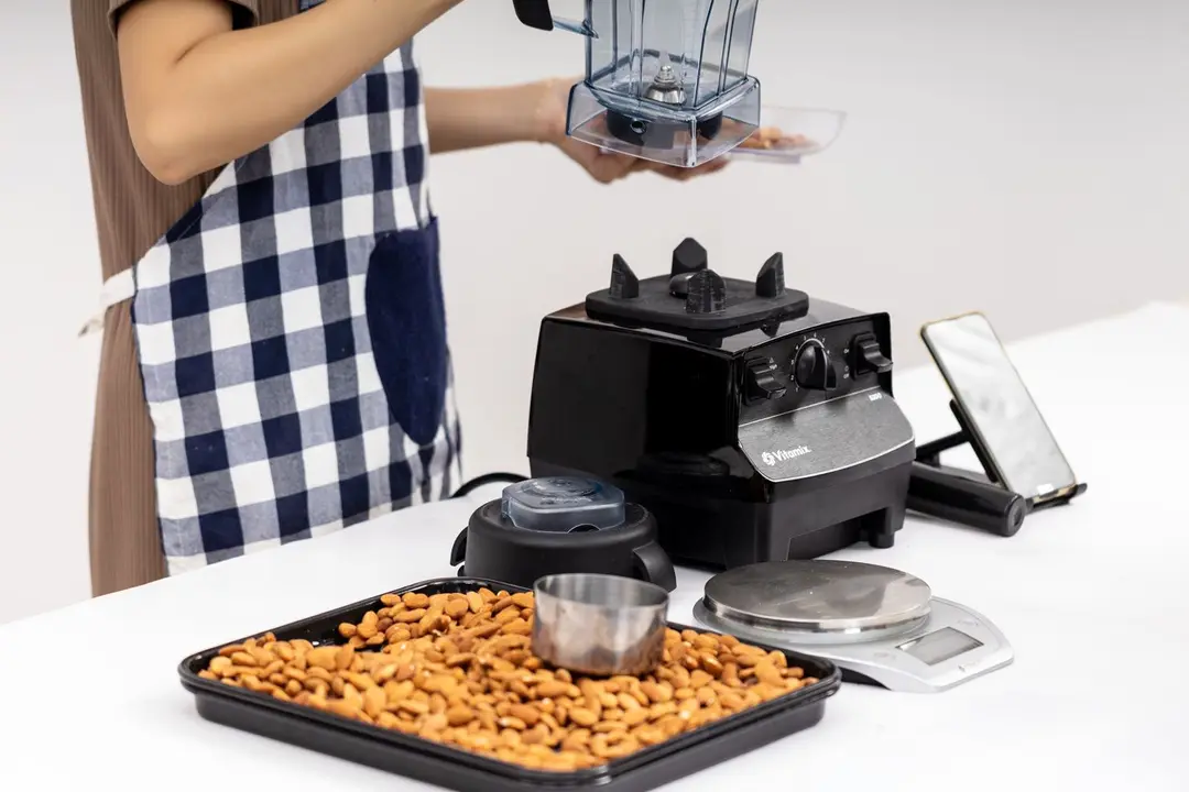 Someone is preparing for the almond butter test with the tray of almonds, a metal cup, and a scale nearby. The Vitamix blender's motor, tamper, and lid are also present, along with a smartphone for timing and recording the blending process.