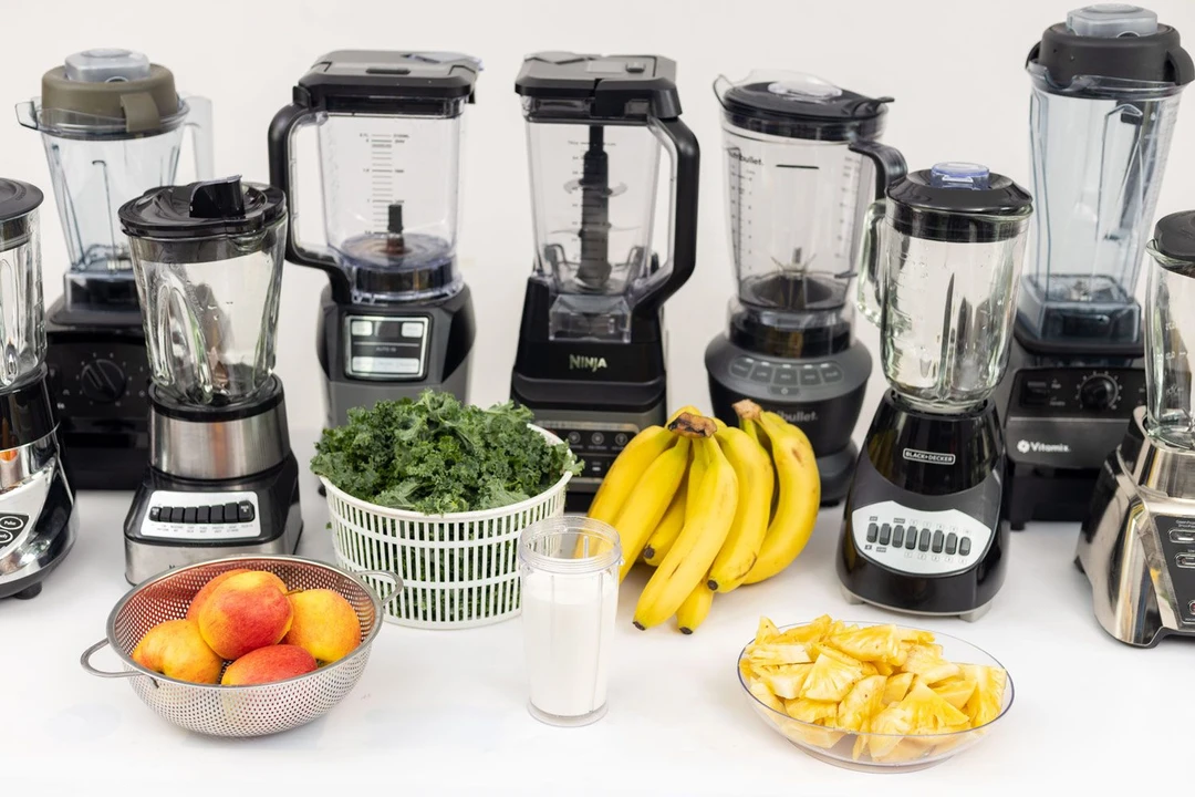 Nine full-sized blenders standing on a table with 5 ingredients for the green smoothie test, including apple, kale, banana, pineapple, and whole milk, next to them.