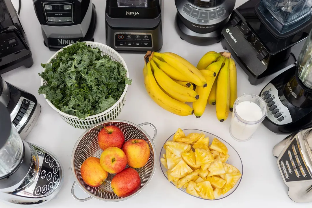 Nine full-sized blenders standing on a table with 5 ingredients for the green smoothie test, including apple, kale, banana, pineapple, and whole milk.