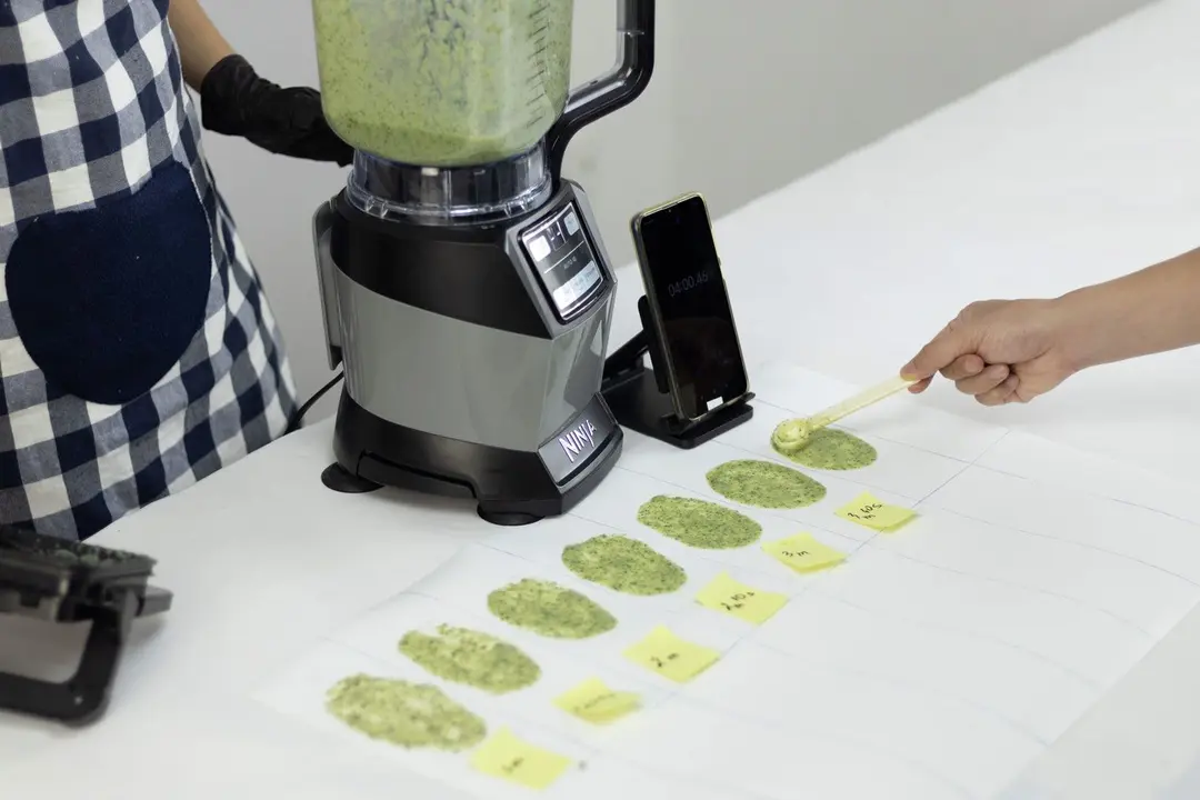 Someone is holding the Ninja AMZ493BRN Compact Kitchen System blender, with a smartphone sitting next to it on a countertop. Another person is using a spoon to spread a small amount of green smoothie onto a white piece of paper.