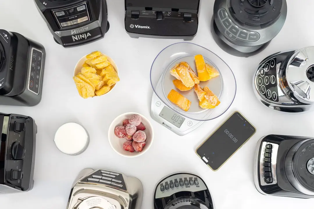 Nine full-sized blenders stand on a table, with three white bowls of testing ingredients (strawberries and pineapples), a cup of milk, a scale containing mangoes, and a smartphone nearby.