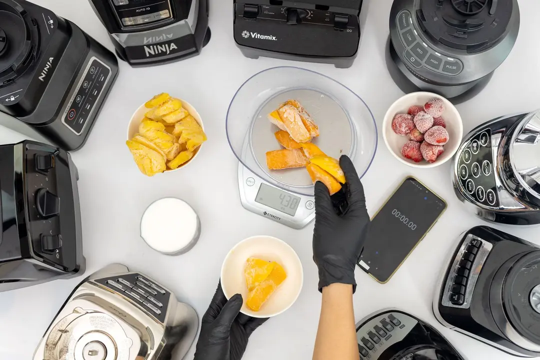 Someone is holding a bowl of mangoes with one hand and using a scale to portion them out with the other hand. Nearby, there are nine full-sized blenders, a cup of milk, bowls of strawberries and pineapple, and a smartphone.