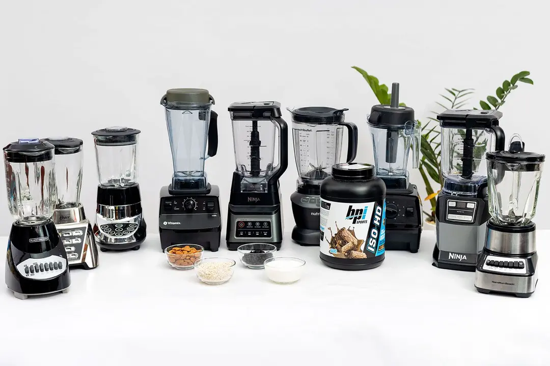 Nine full-sized blenders sit on a table surrounded by four bowls containing testing ingredients (almonds, oatmeal, dried berries, and milk) and a container of protein powder.