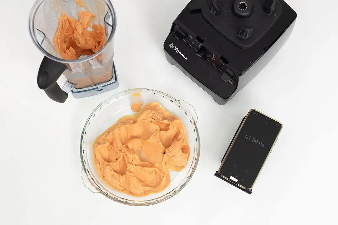 The Vitamix 5200's motor base, its container with a portion of the smoothie, a bowl of the completed smoothie, and a smartphone are on a table.