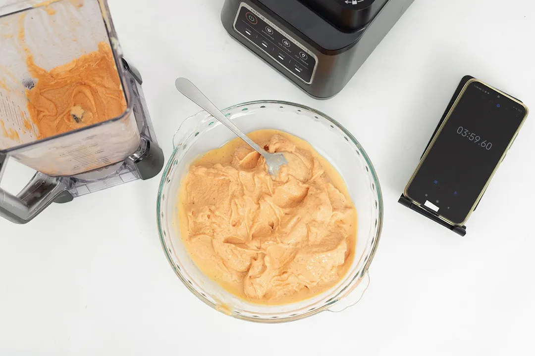 The Ninja BN401's motor base, its container with a portion of the smoothie, a bowl of the completed smoothie, and a smartphone are on a table. 