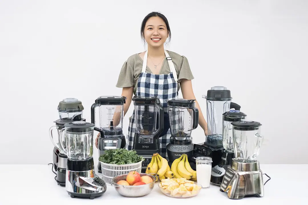 A woman stands beside a table with nine full-sized blenders and an array of fresh ingredients (apple, kale, banana, pineapple, and whole milk), ready for the smoothie test.