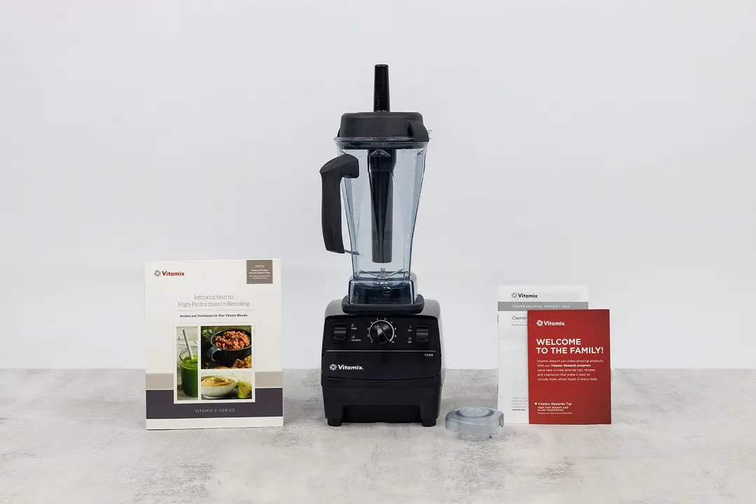The Vitamix 5200 blender stands on a table, with a recipe booklet, a lid plug, and a user manual by its sides.