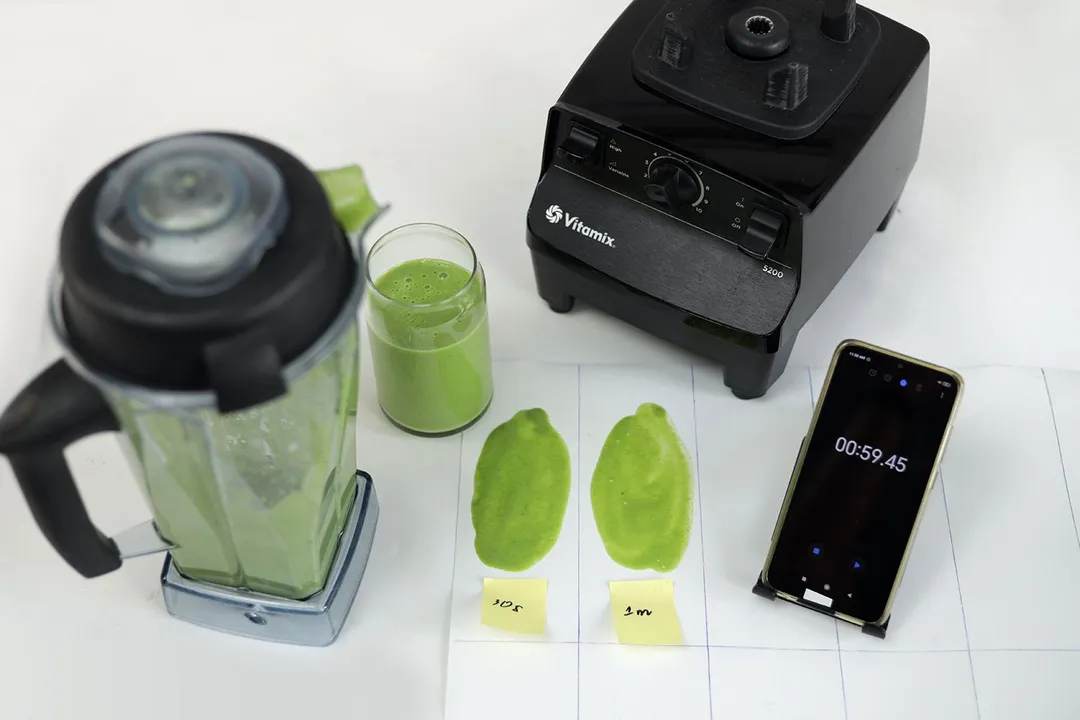 The Vitamix 5200 motor base and its container stand on a table, accompanied by a glass filled with a green smoothie. Next to it, a white paper features two distinct smoothies spread while a smartphone showcases the blending time of 59 seconds.