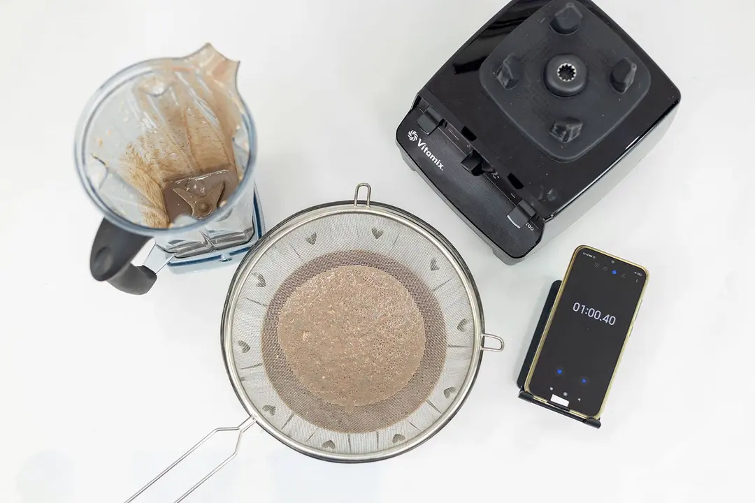 The Vitamix 5200 motor base stands beside the container. Next to it, a metal mesh strainer filtered its protein shake while a smartphone displayed a blending time of 1 minute.