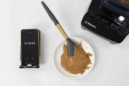 The Vitamix 5200 is beside a white plate containing almond butter with a spatula nestled inside, and a smartphone revealing a blending time of 1 minute.