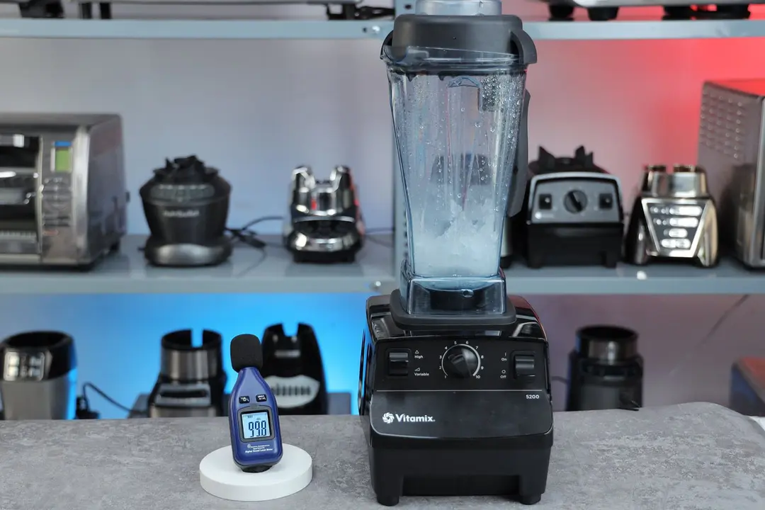 The Vitamix 5200 blender is on a table, accompanied by a sound level meter displaying its noise level nearby. In the background, other blenders that we tested sit on a nearby shelf.