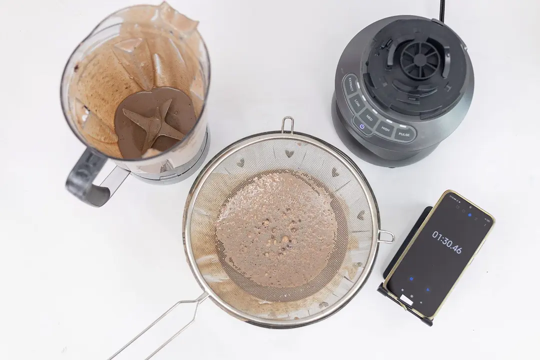 The NutriBullet motor base stands beside the container. Next to it, a metal mesh strainer filtered its protein shake while a smartphone displayed a blending time of 1 minute 30 second.