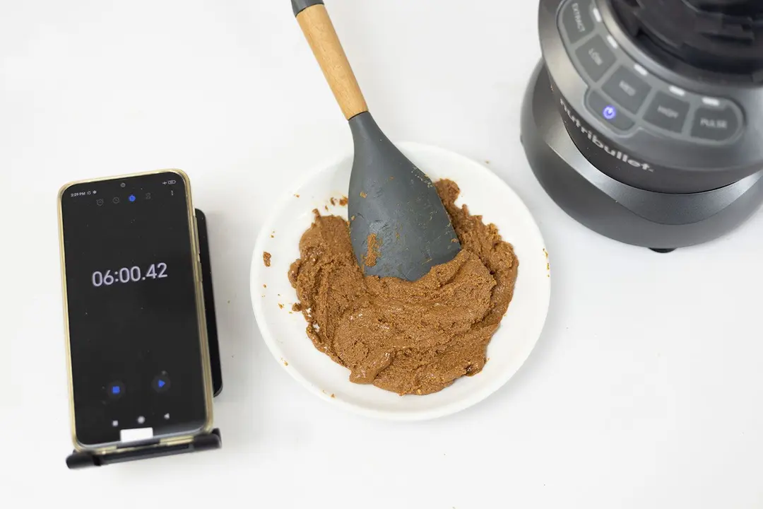 The NutriBullet is beside a white plate containing almond butter with a spatula nestled inside, and a smartphone revealing a blending time of 6 minutes.