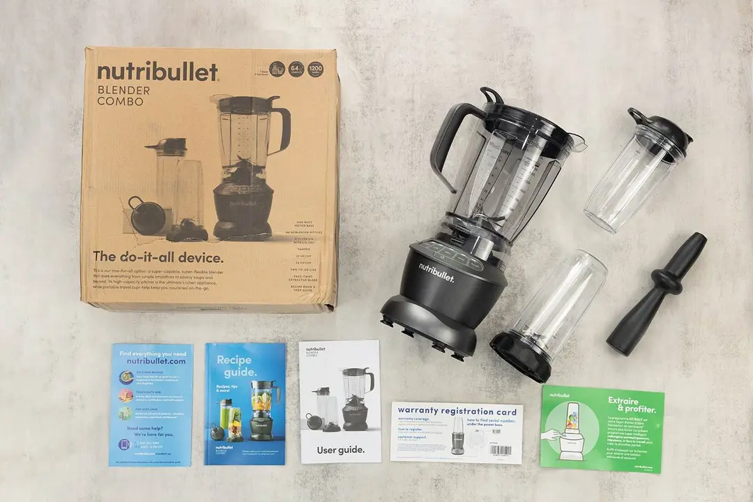 A display of the NutriBullet ZNBF30500Z blender and its accessories, including the tamper, carton box, recipe booklet, user manual, extra blending pitcher, and handled cup, on a table.