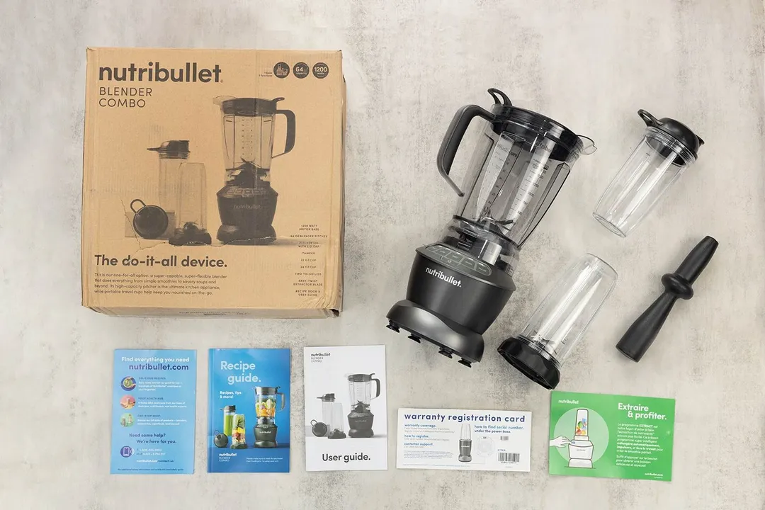 A display of the NutriBullet ZNBF30500Z blender and its accessories, including the tamper, carton box, recipe booklet, user manual, extra blending pitcher, and handled cup, on a table.